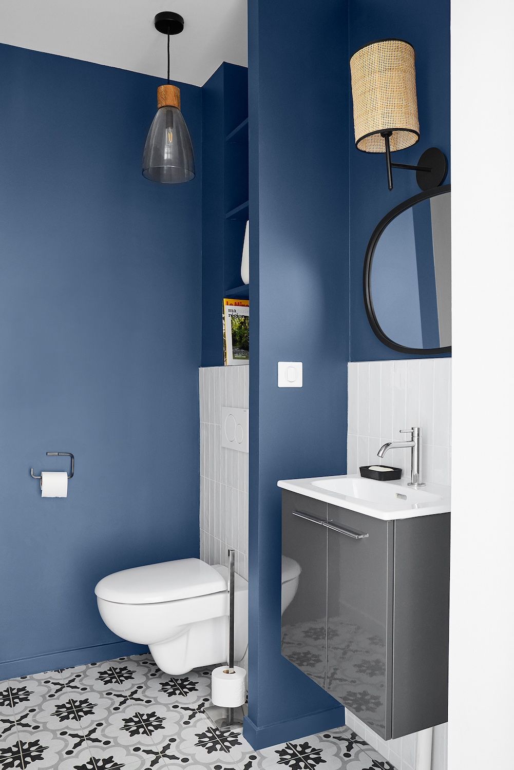 Salle de bains bleue avec toilettes, lavabo suspendu, miroir rond, carrelage à motifs au sol et suspension lumineuse.