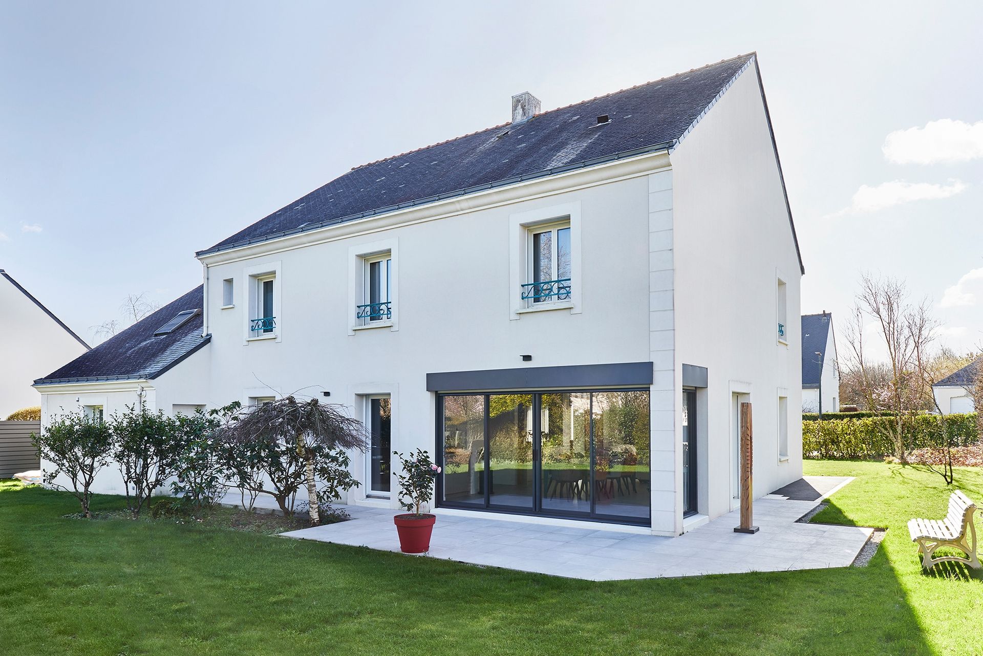 Maison blanche à deux étages avec toit sombre, grandes fenêtres et pelouse verte baignée de lumière naturelle.