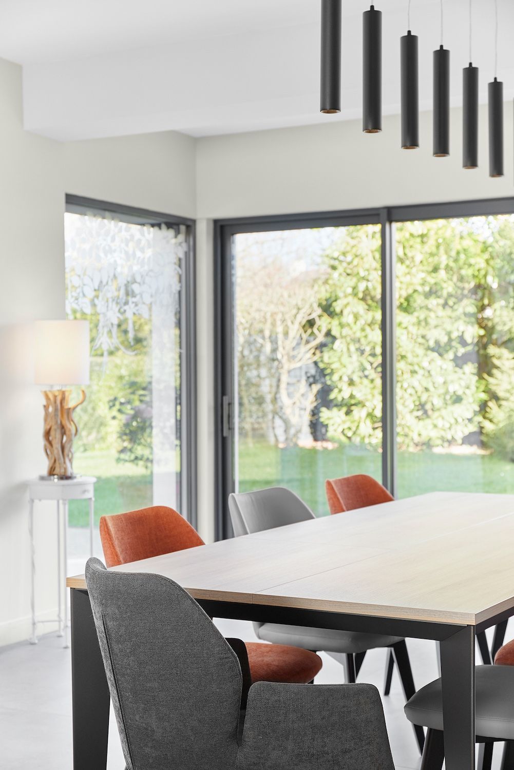 Salle à manger moderne avec de grandes fenêtres, une table blanche et des suspensions noires donnant sur un jardin verdoyant