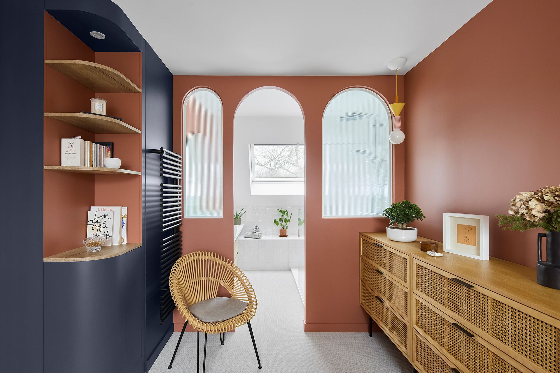 Couloir moderne avec murs en terre cuite, miroirs arqués, fauteuil en osier et console en rotin avec plantes.