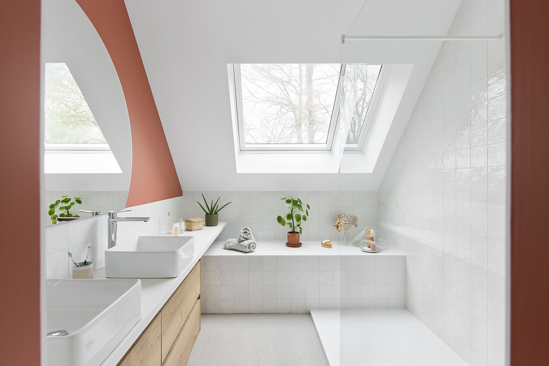 Salle de bain mansardée d'un blanc éclatant avec puits de lumière, baignoire, lavabo et petites plantes en pot sur une étagère.