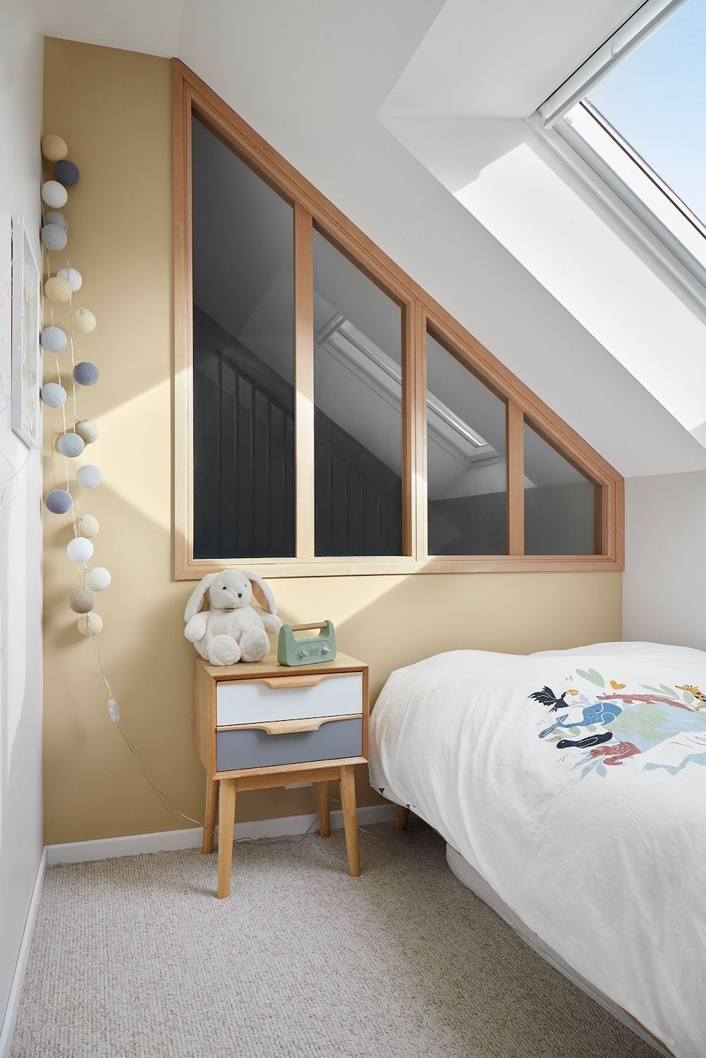 Chambre mansardée confortable avec puits de lumière incliné, fenêtres à cadre en bois, table de chevet et couvre-lit blanc.