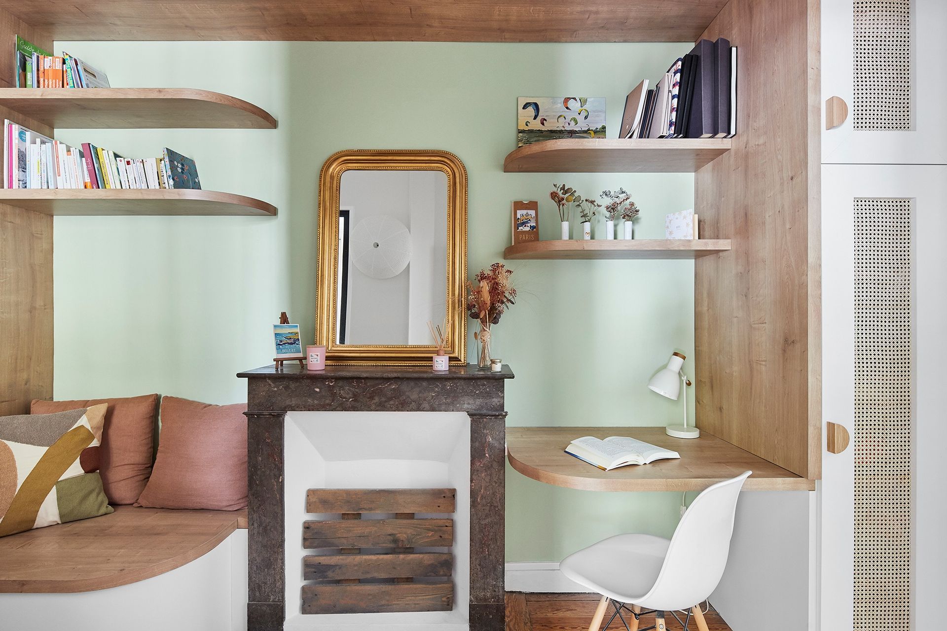 Coin lecture confortable avec fenêtre intégrée, étagères, coussins roses, petit bureau et cheminée avec miroir.