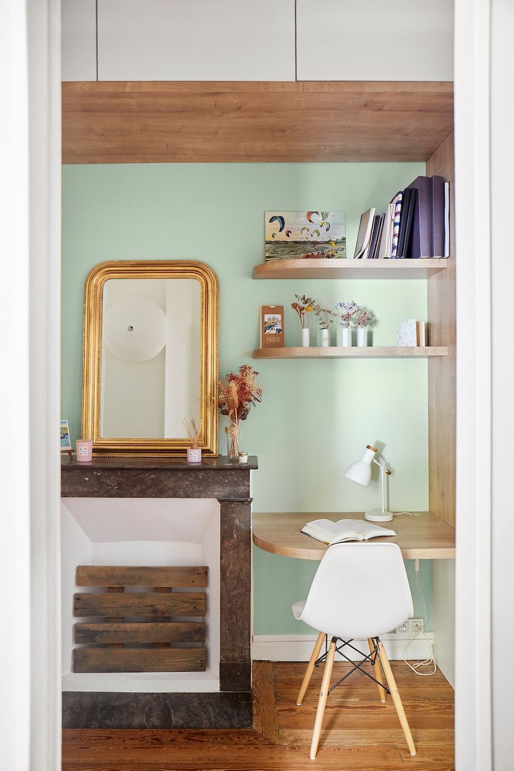Petit bureau à domicile aux tons pastel, avec miroir doré, étagères et chaise blanche près de la cheminée.