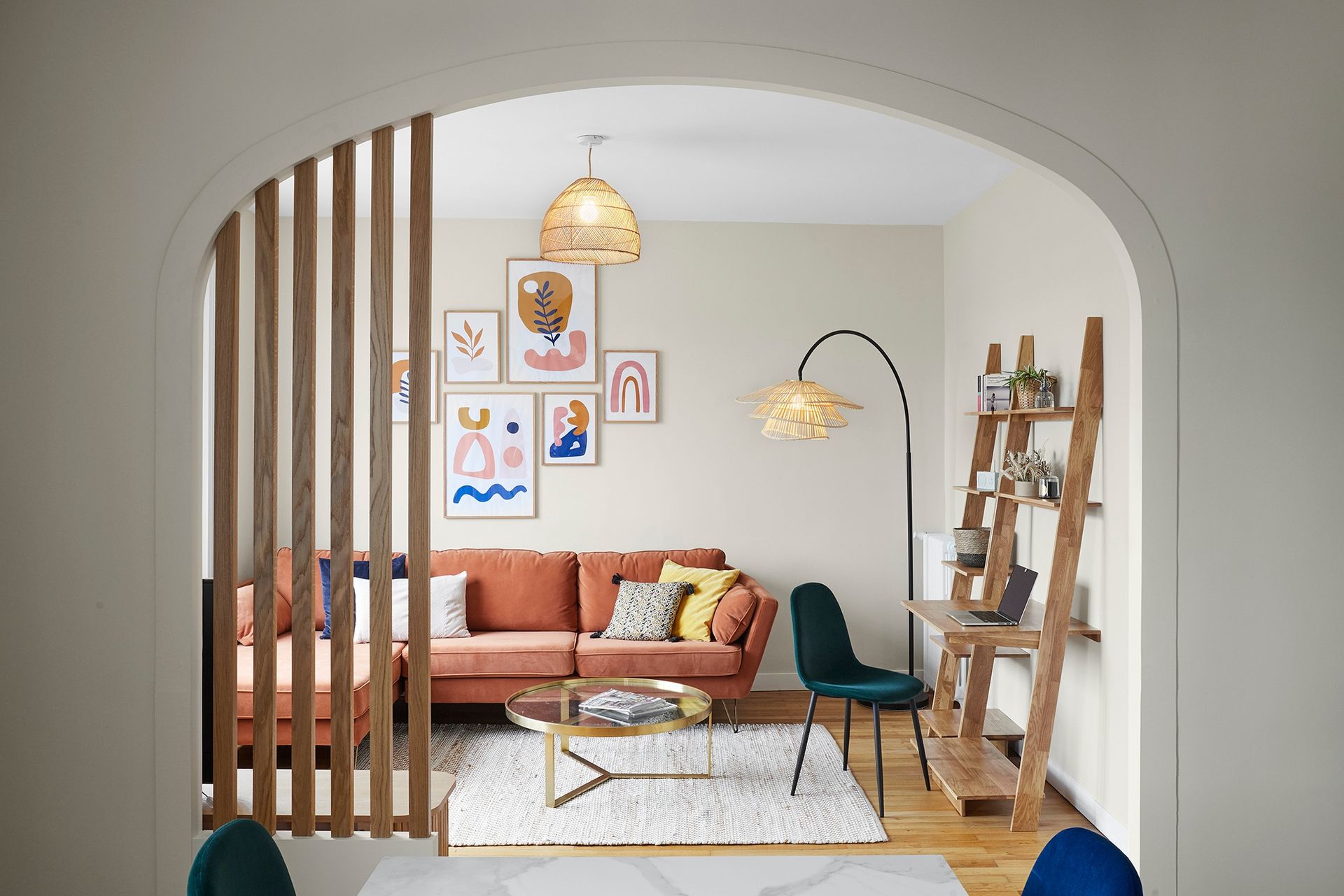 Salon moderne avec un canapé pêche, des tableaux encadrés, des suspensions lumineuses et une étagère échelle, vu à travers une porte cintrée.