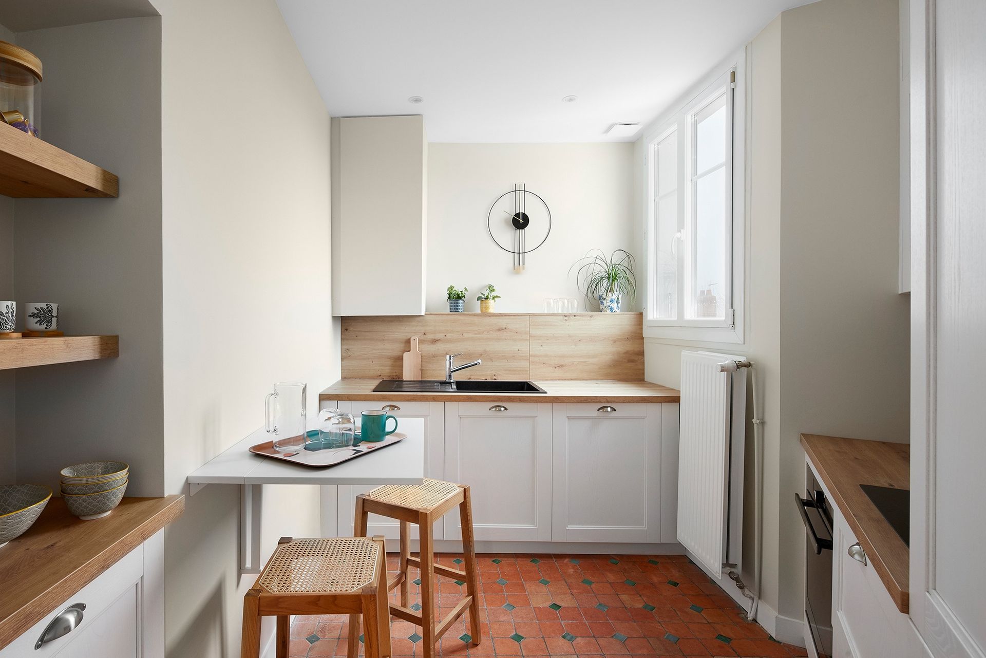 Cuisine étroite et lumineuse avec placards blancs, plans de travail en bois, sol en carrelage de terre cuite et une petite table près de la fenêtre.