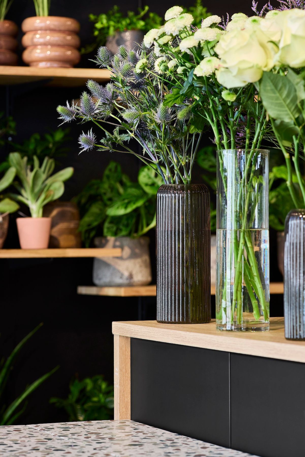 Des compositions florales dans de grands vases en verre, posées sur un comptoir en bois dans une jardinerie.