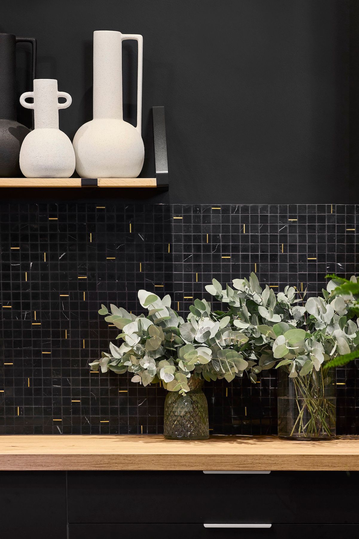 Étagère de cuisine noire avec vases blancs, crédence en carrelage noir et fleurs vertes sur un comptoir en bois.