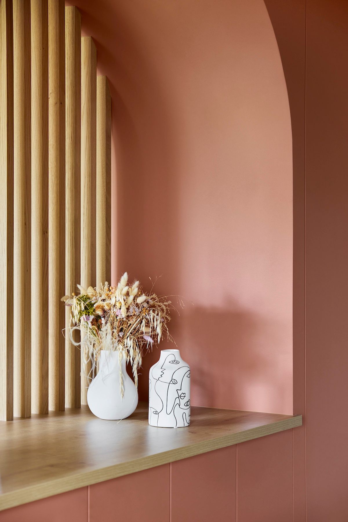 Étagère rose minimaliste avec vases blancs et fleurs séchées à côté de lattes de bois verticales.