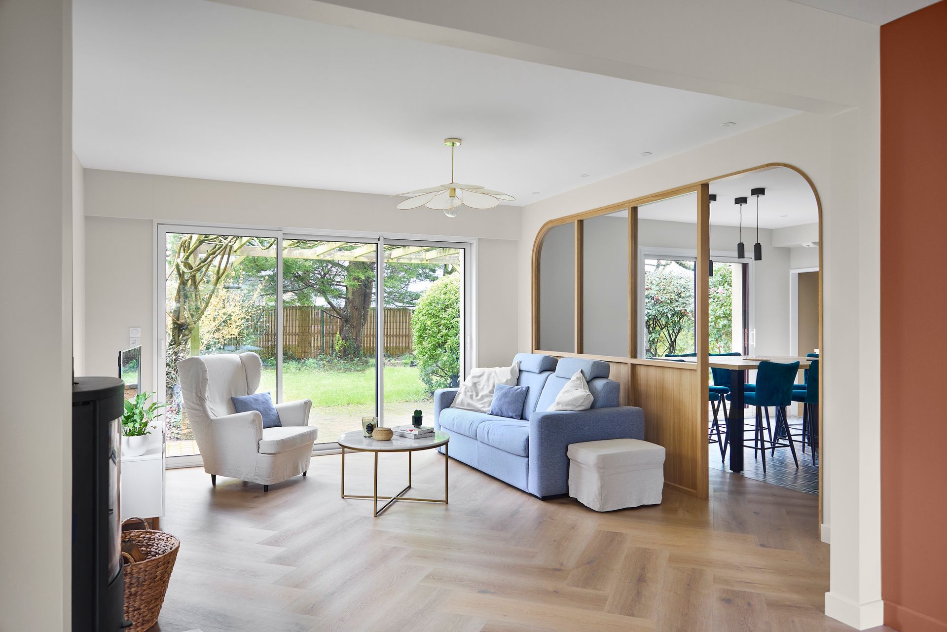 Salon lumineux avec canapé bleu, fauteuil blanc, table basse et grandes portes coulissantes donnant sur un jardin