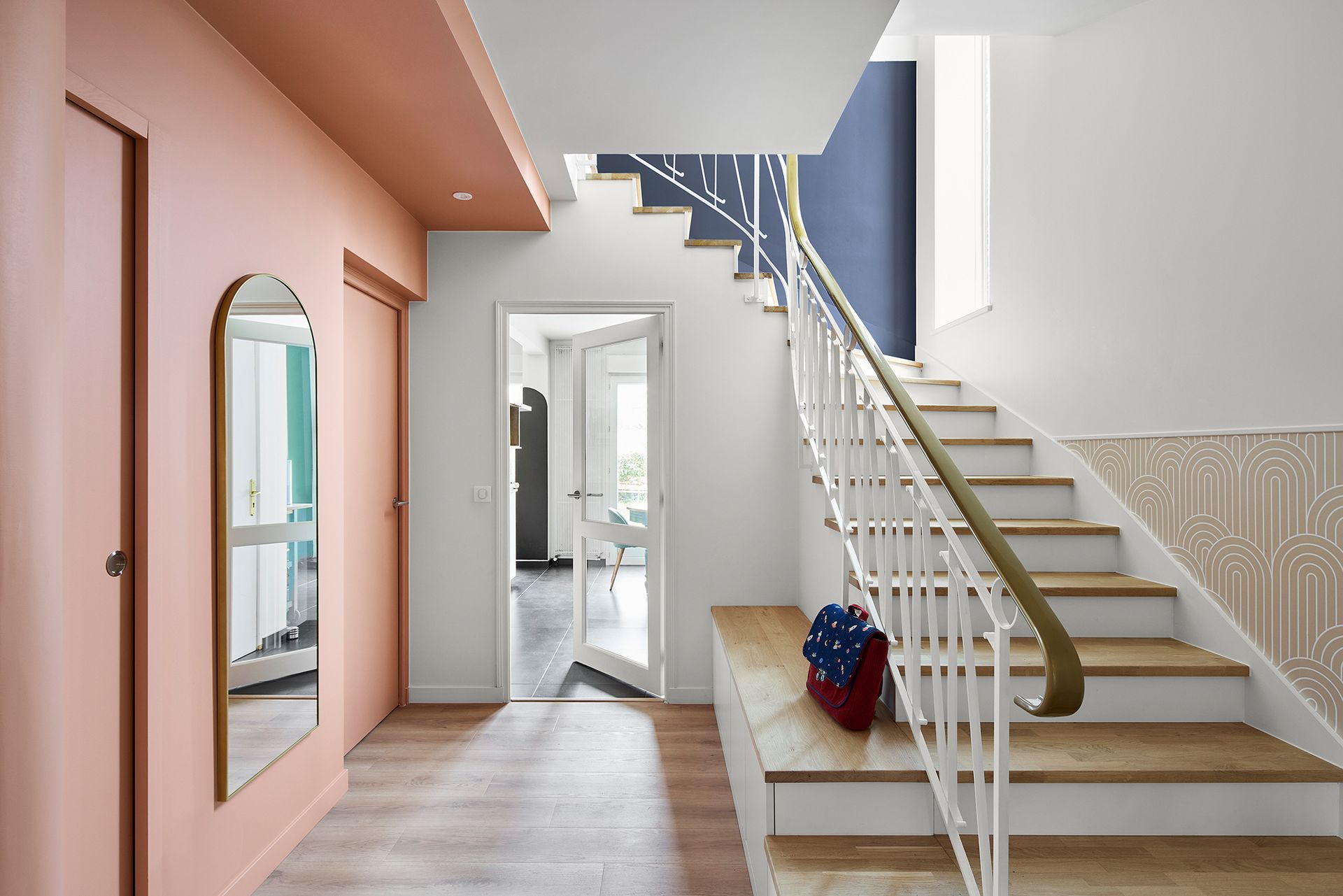 Entrée moderne avec escalier, mur couleur pêche, miroir arqué et porte ouverte sur une pièce lumineuse