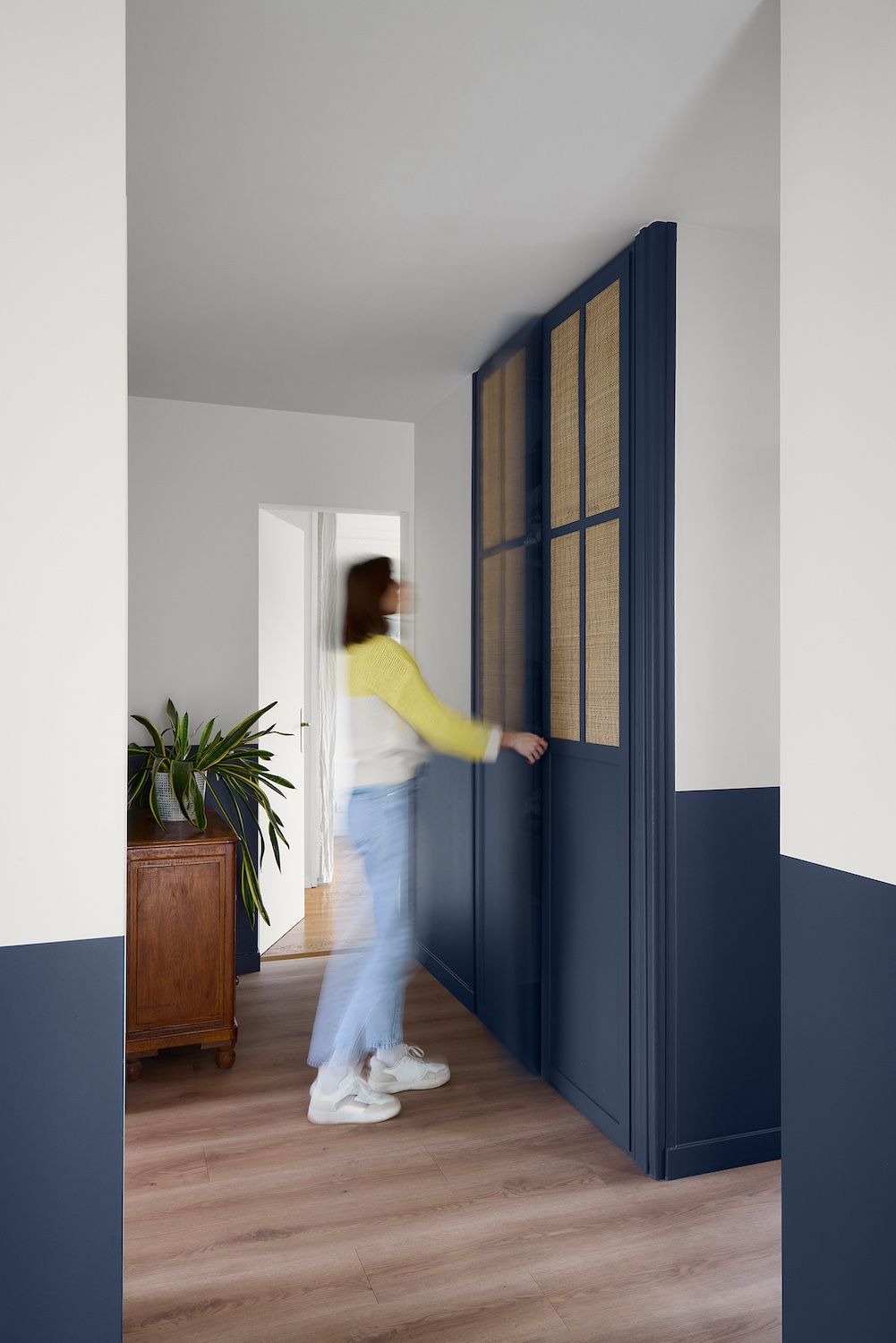 Une personne ouvre une armoire bleu marine à portes tressées dans un couloir blanc au parquet.