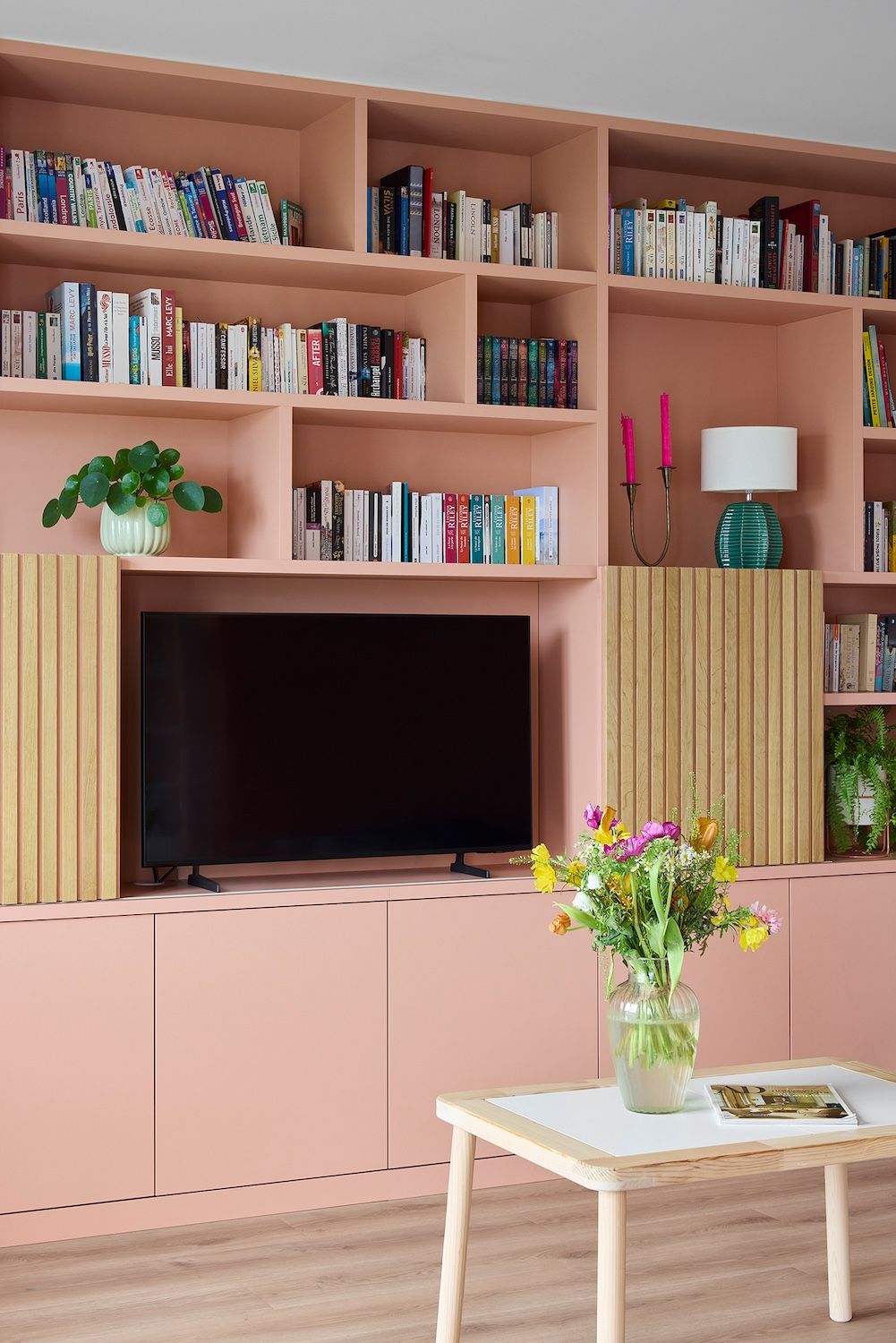 Dans un salon, des étagères roses encastrées accueillent des livres, une télévision, des plantes et une table basse ornée de fleurs.
