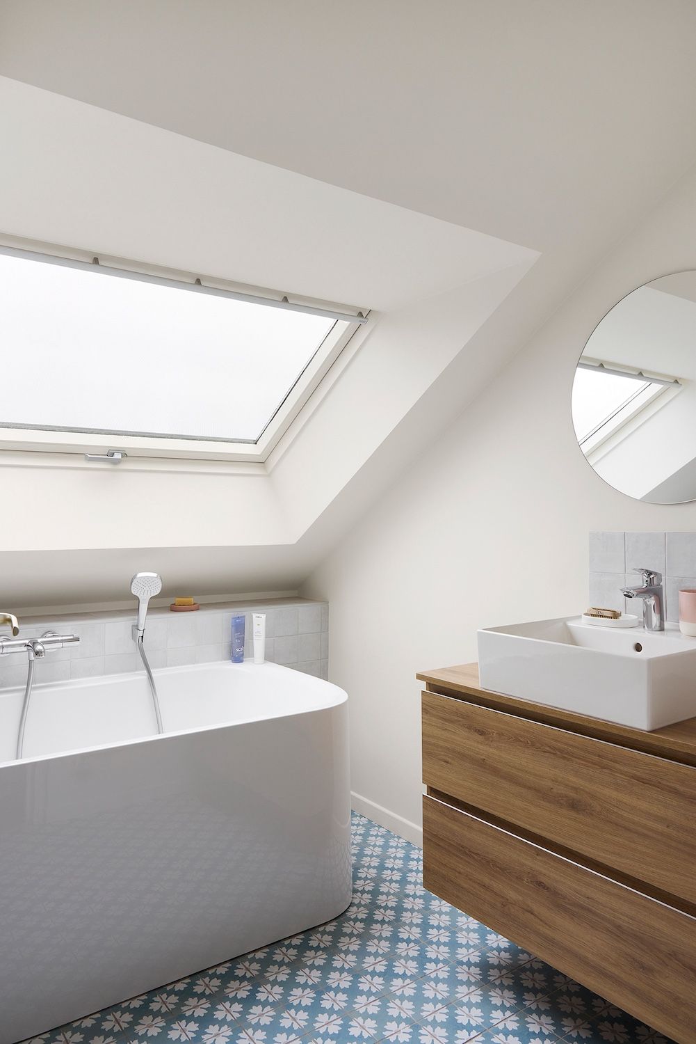 Salle de bains moderne avec puits de lumière, baignoire îlot, meuble-lavabo en bois, miroir rond et carrelage au sol bleu à motifs.