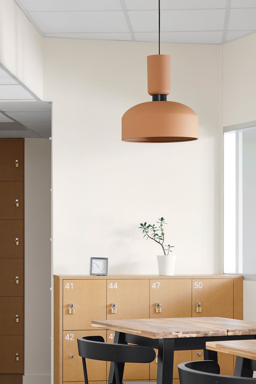 Salle à manger moderne avec tables en bois, chaises noires, placards beiges et une grande suspension lumineuse orange.