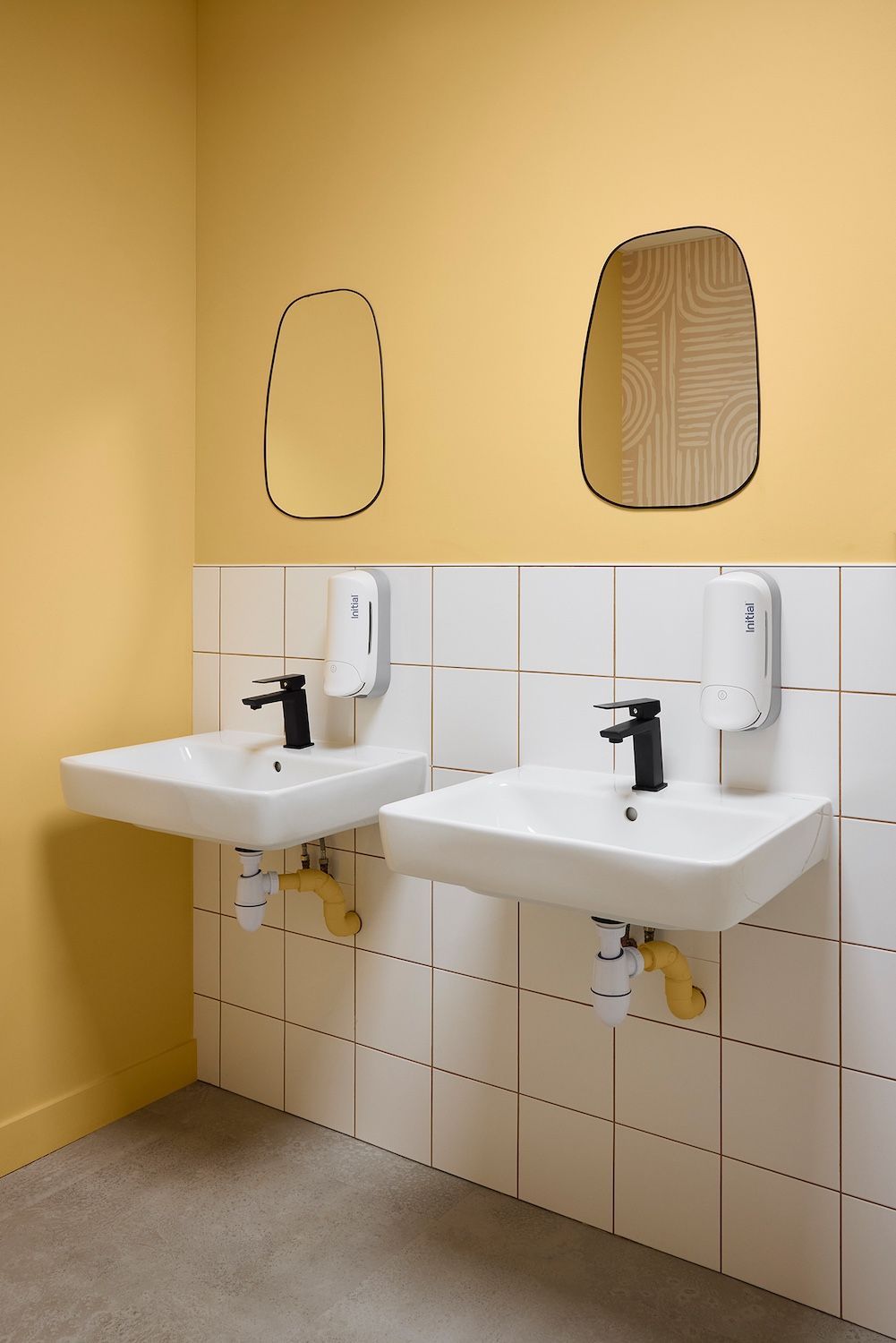 Deux lavabos blancs avec robinets noirs dans des toilettes jaunes, avec des miroirs au-dessus et des murs inférieurs carrelés.