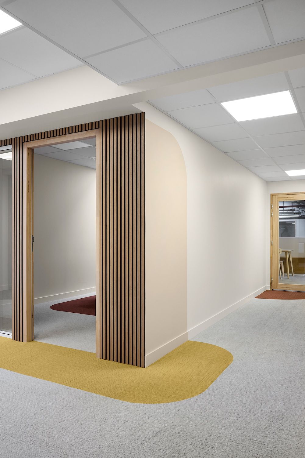 Couloir de bureau moderne avec porte à lattes de bois, murs blancs, moquette grise et accents jaunes au sol