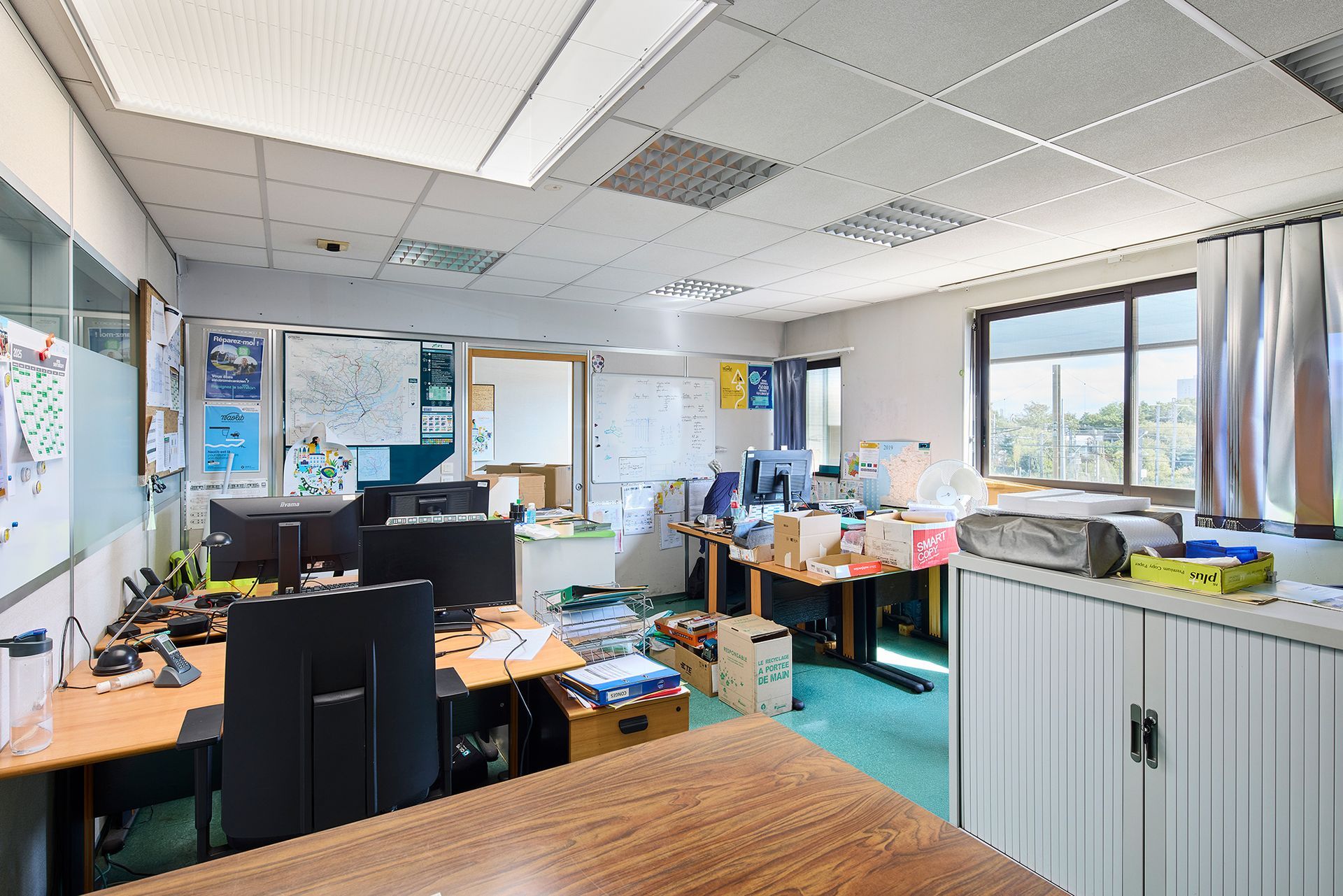 Bureau encombré de bureaux, d'ordinateurs, de papiers et de grandes fenêtres laissant entrer la lumière du jour