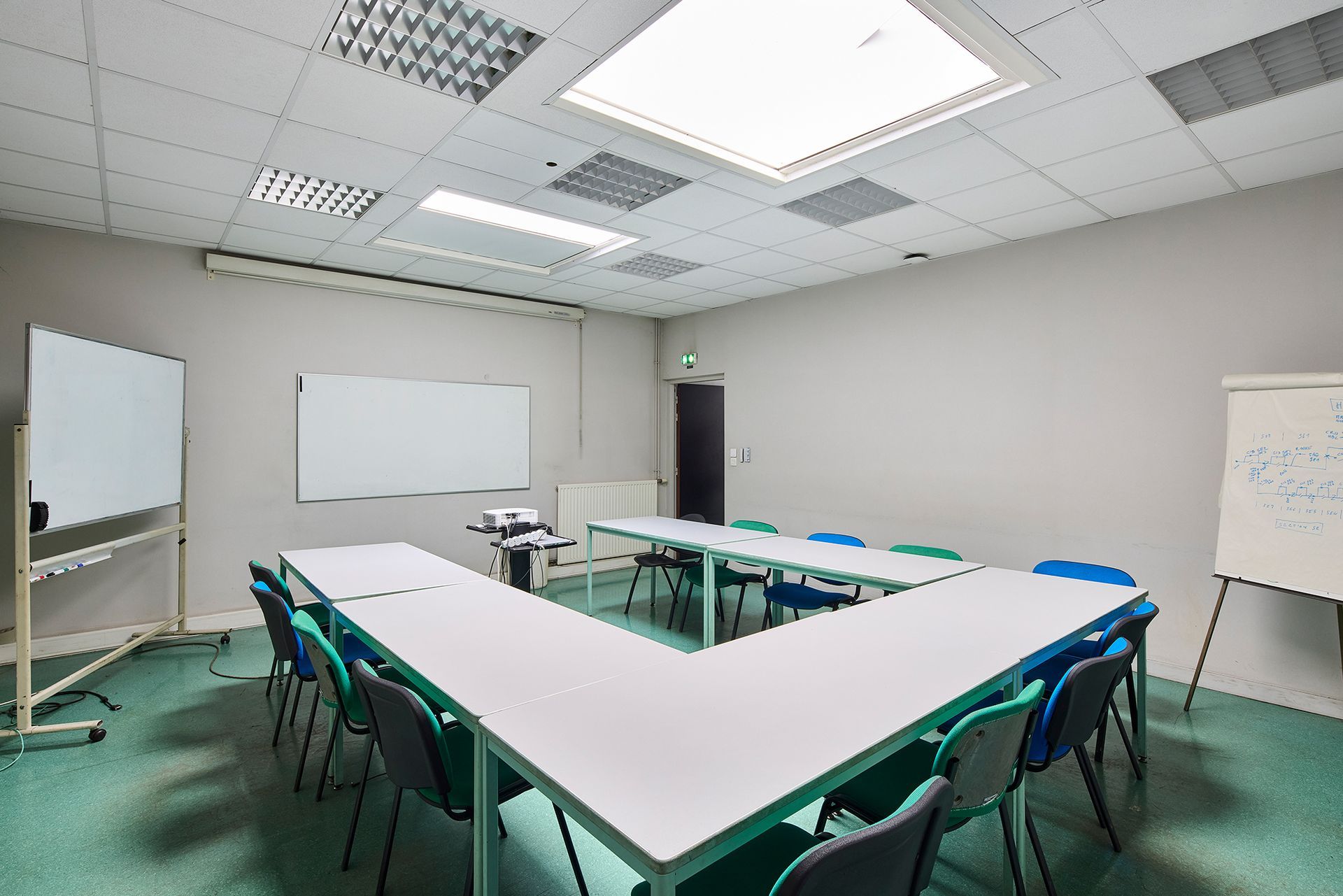 Salle de classe vide avec tables en forme de U, chaises vertes, tableaux blancs et plafonniers