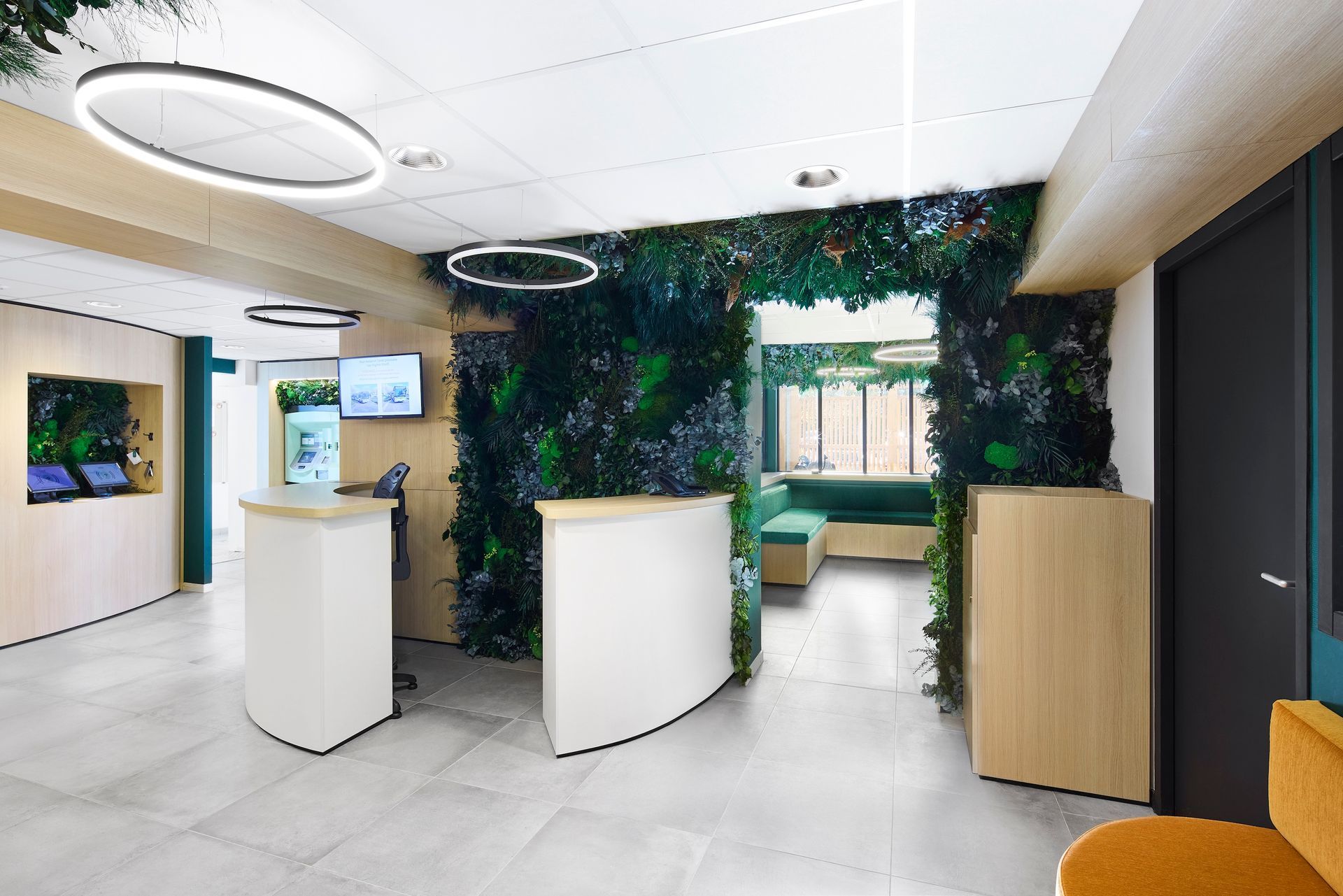 Espace d'accueil moderne avec bureaux blancs, éléments en bois et mur végétal près d'une fenêtre.