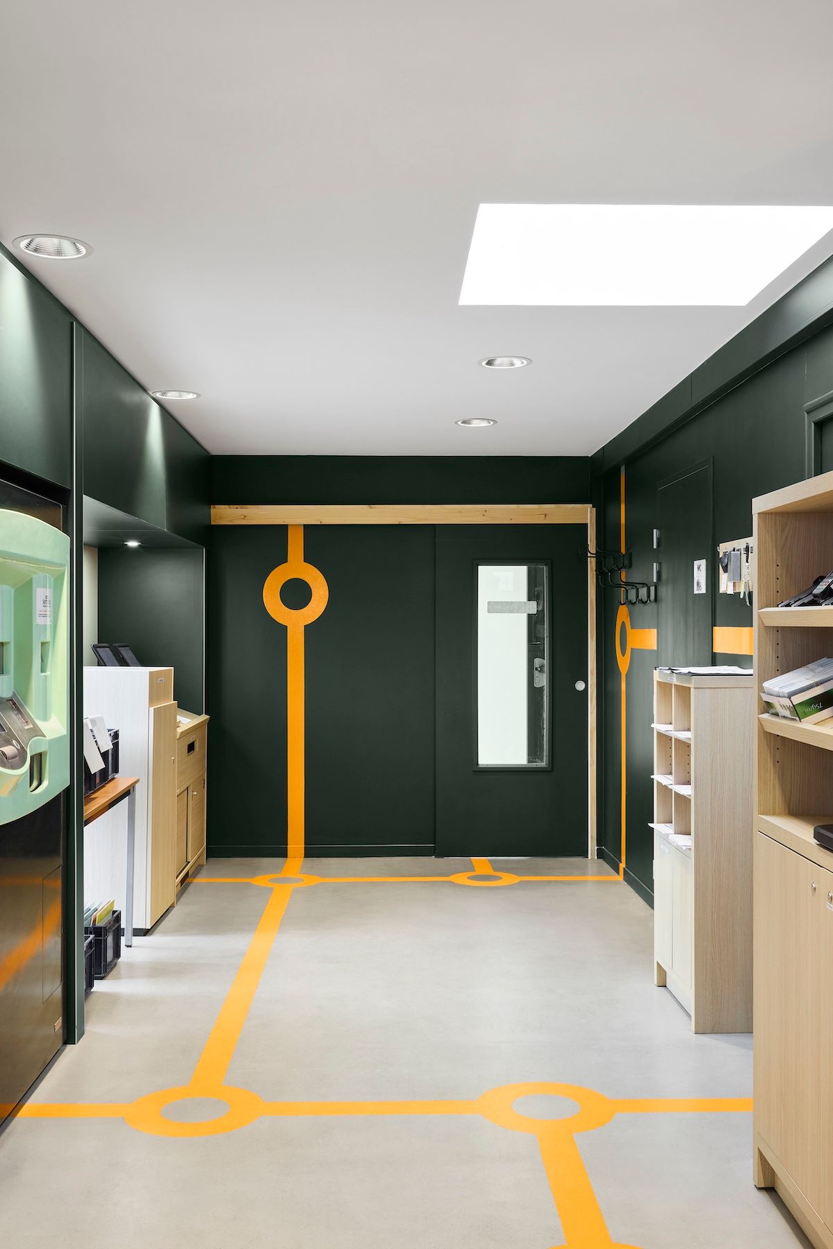 Couloir moderne avec marquages ​​au sol orange, murs vert foncé, puits de lumière et armoires de rangement en bois.