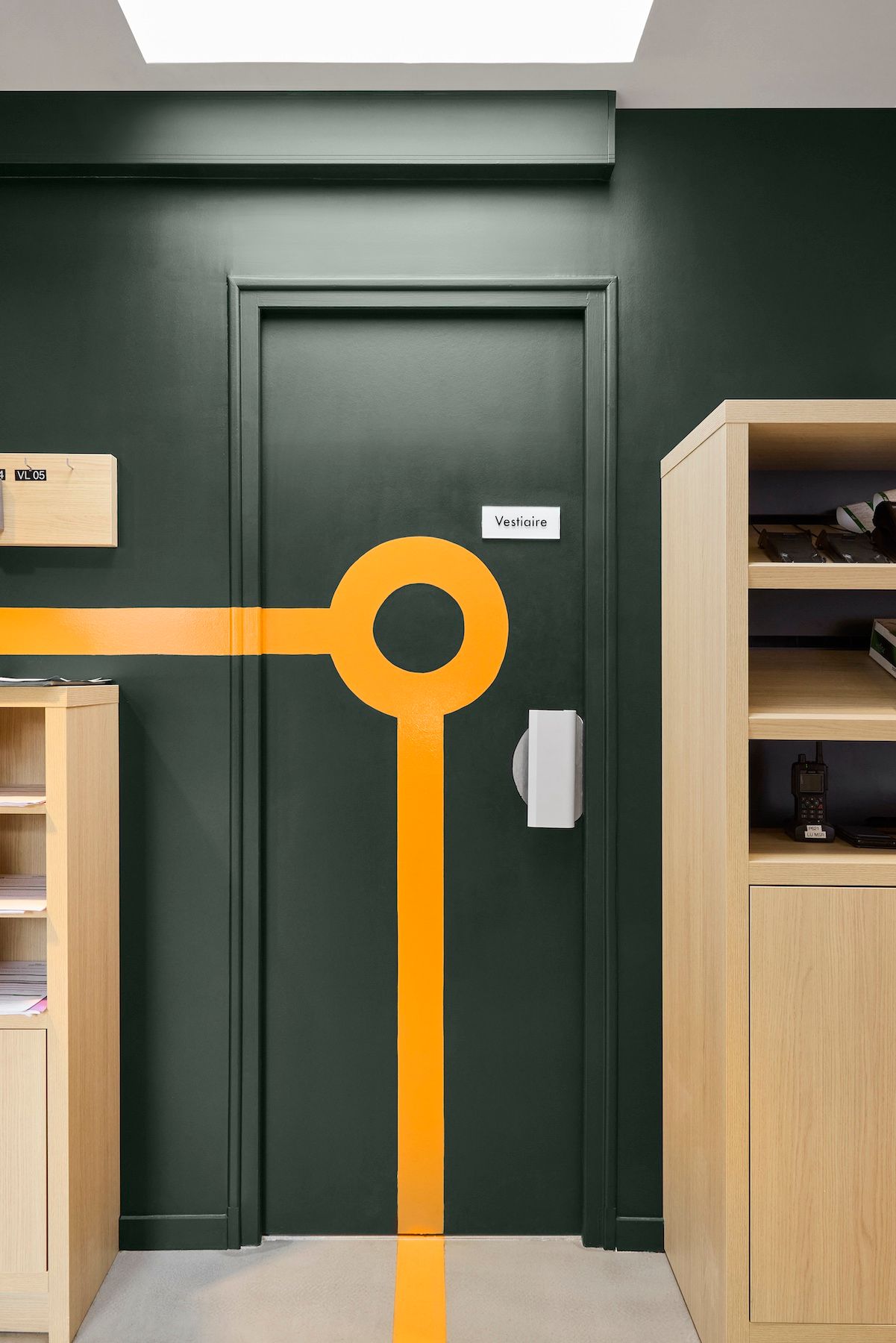 Porte vert foncé ornée d'une ligne et d'un cercle orange dans un couloir moderne, encadrée par des étagères en bois.