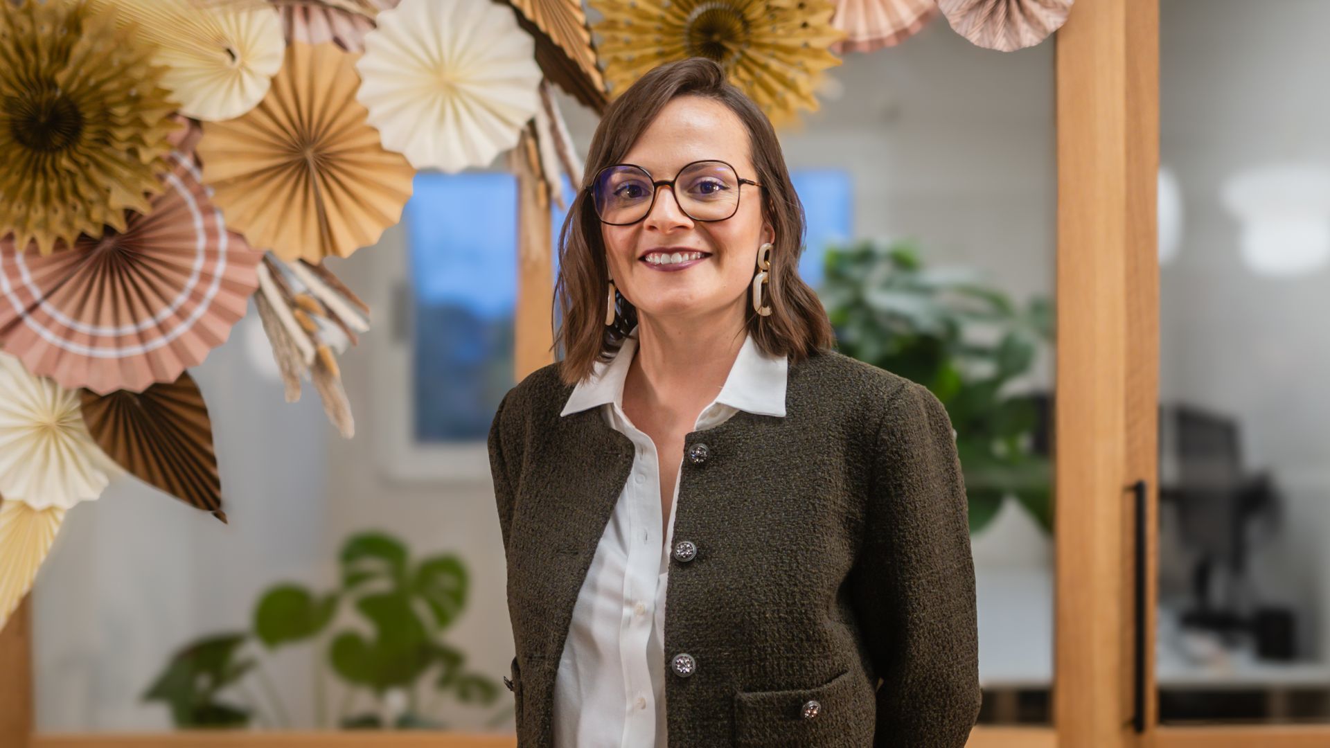 Femme souriante en blazer, debout à l'intérieur à côté de fleurs décoratives en papier.