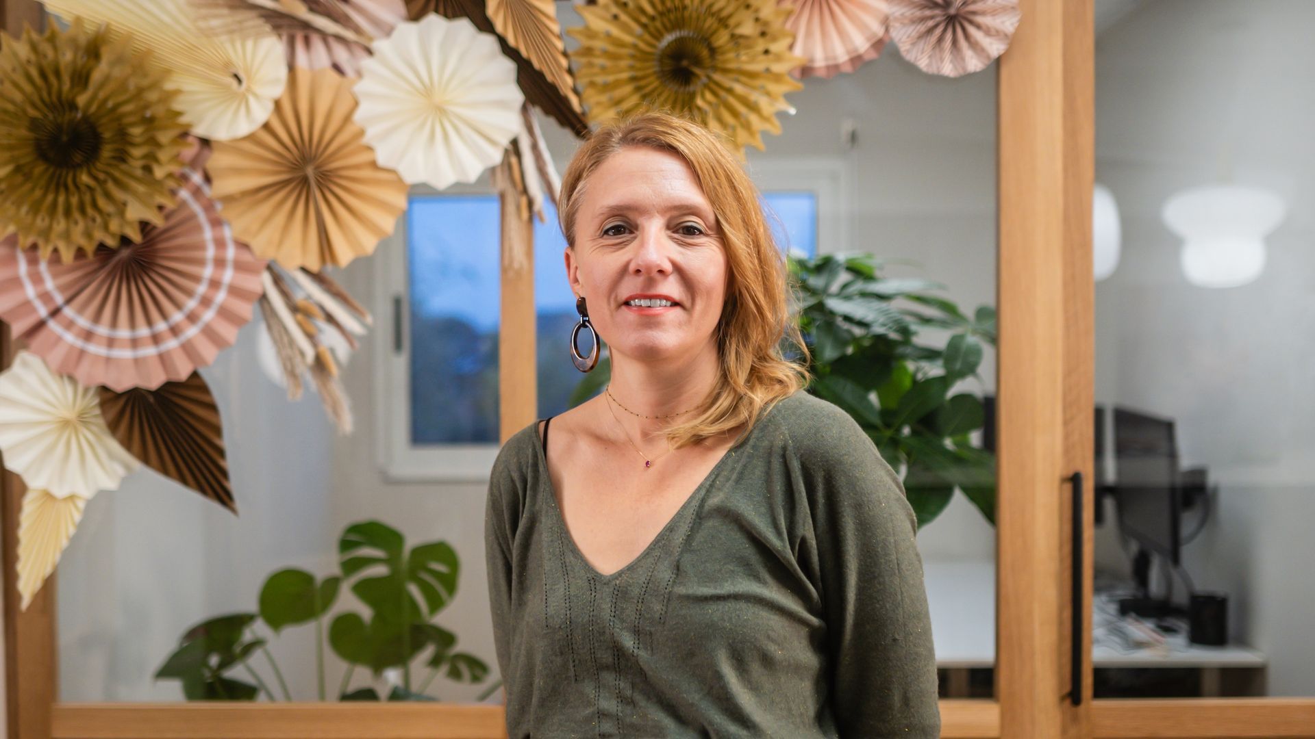 Une femme souriante, vêtue d'un pull vert, se tient à l'intérieur, à côté d'un mur décoratif de fleurs en papier.