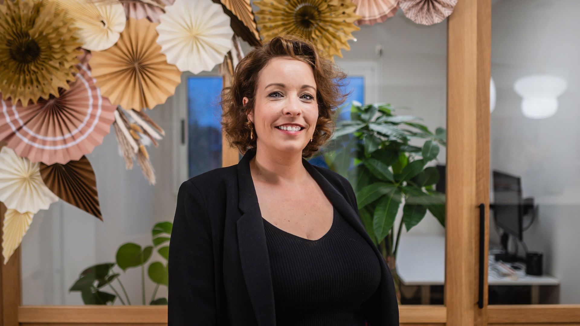 Une femme souriante, vêtue d'un blazer noir, se tient dans un bureau, des fleurs décoratives en papier derrière elle.