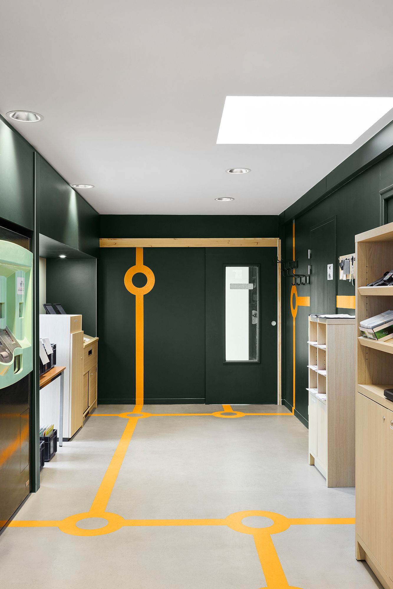 Couloir moderne avec murs vert foncé, étagères en bois et marquages ​​au sol orange menant à une porte vitrée.