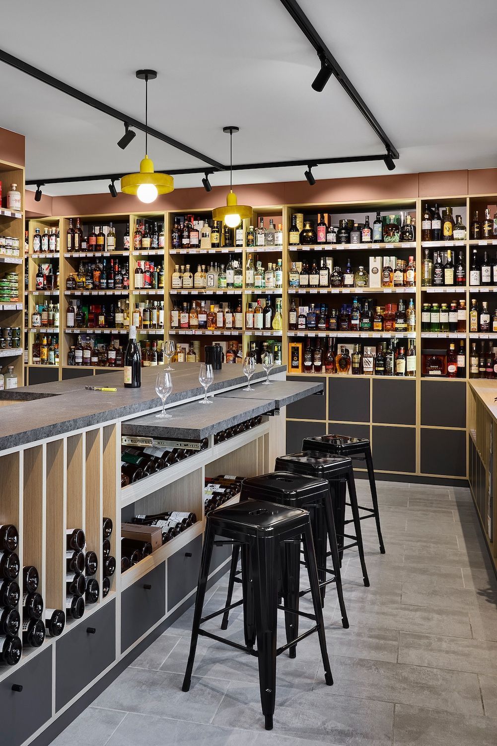 Intérieur de bar à vin avec étagères murales remplies de bouteilles, tabourets noirs et luminaires suspendus jaunes.