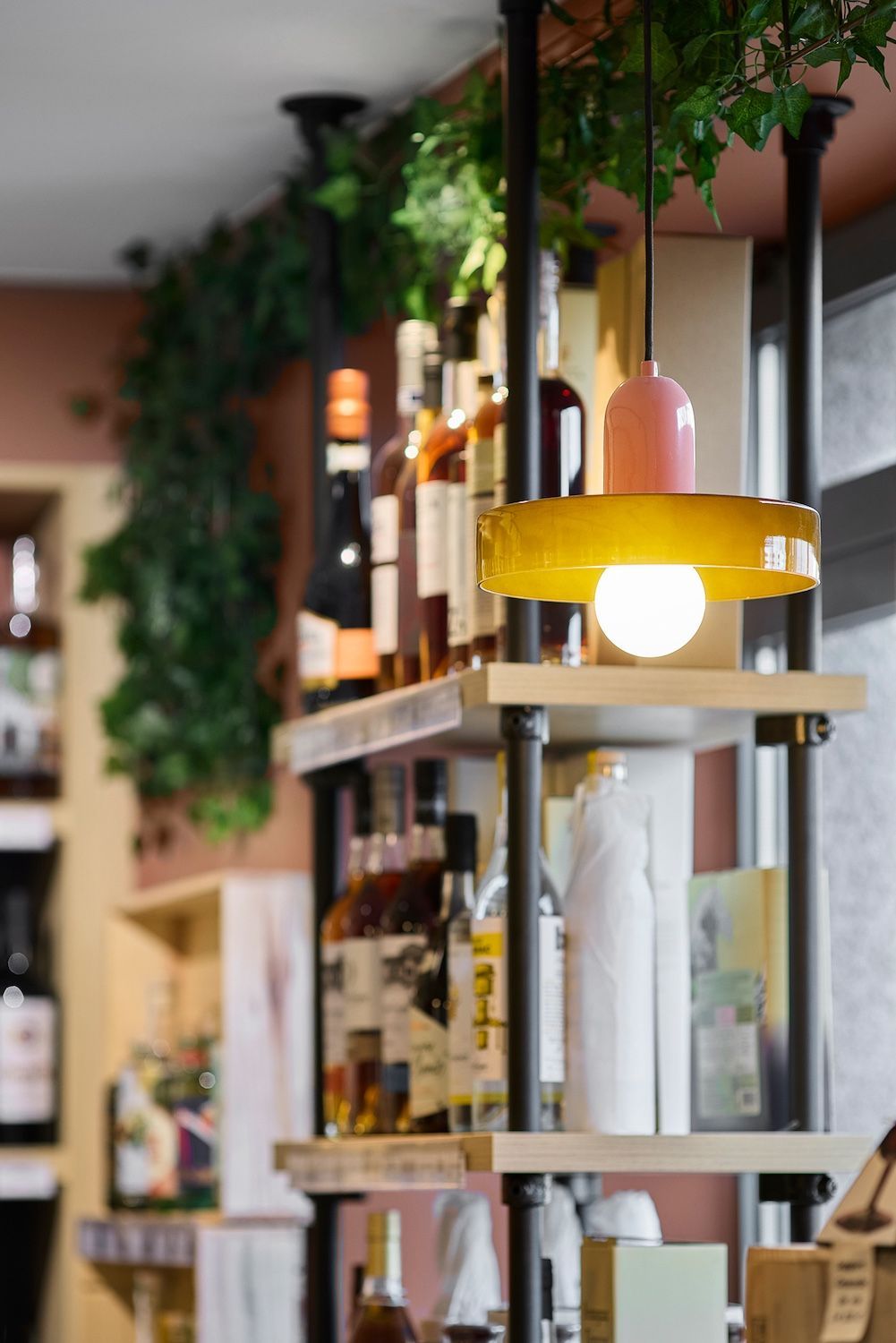 Étagère en bois avec bouteilles et objets de décoration dans un café cosy, éclairé par une lampe suspendue.
