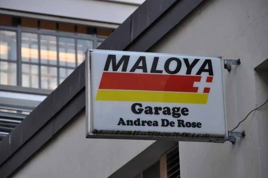 Enseigne du garage MALOYA avec des rayures noires, rouges et dorées, portant l'inscription Andrea De Rose sur un bâtiment
