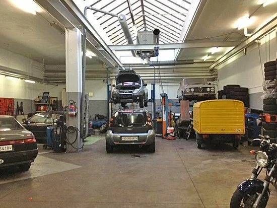 Atelier de réparation automobile avec plusieurs voitures, un camion jaune et une moto à l'intérieur d'un grand garage