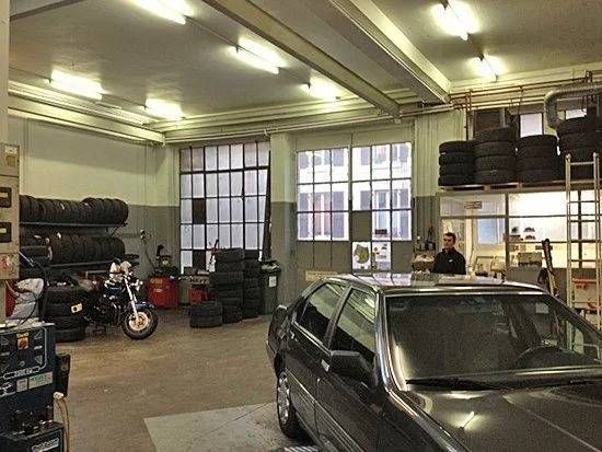 Une voiture dans un garage encombré, avec des étagères, des outils et une moto sous des néons.