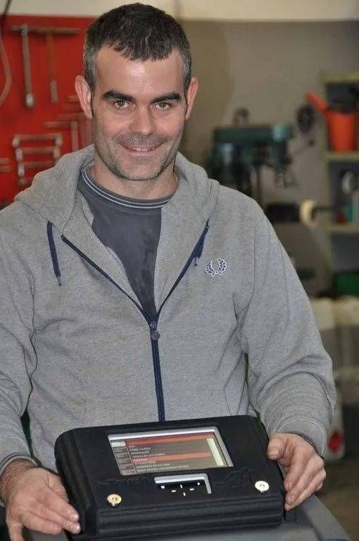 Un homme tient un appareil électronique noir dans un atelier, avec des armoires à outils rouges en arrière-plan.