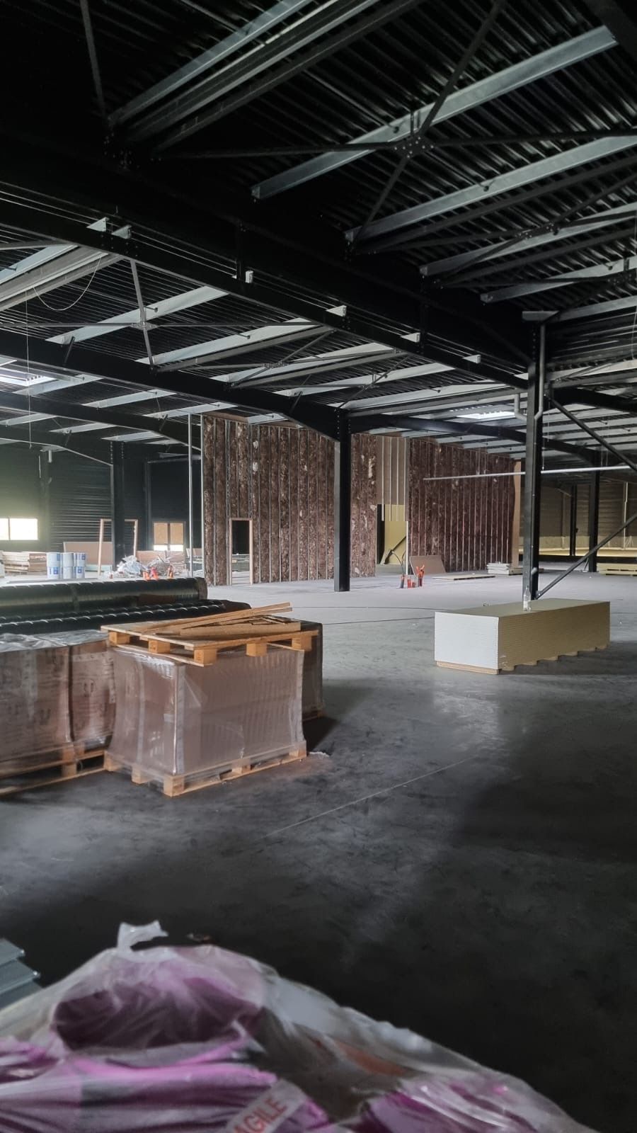 Intérieur industriel vide avec poutres apparentes au plafond, sol en béton et matériaux de construction empilés.