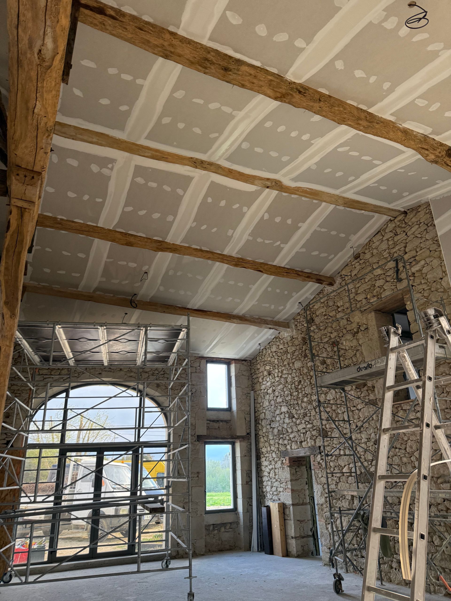 Intérieur en pierre avec fenêtre cintrée, poutres apparentes au plafond et échelle adossée au mur.