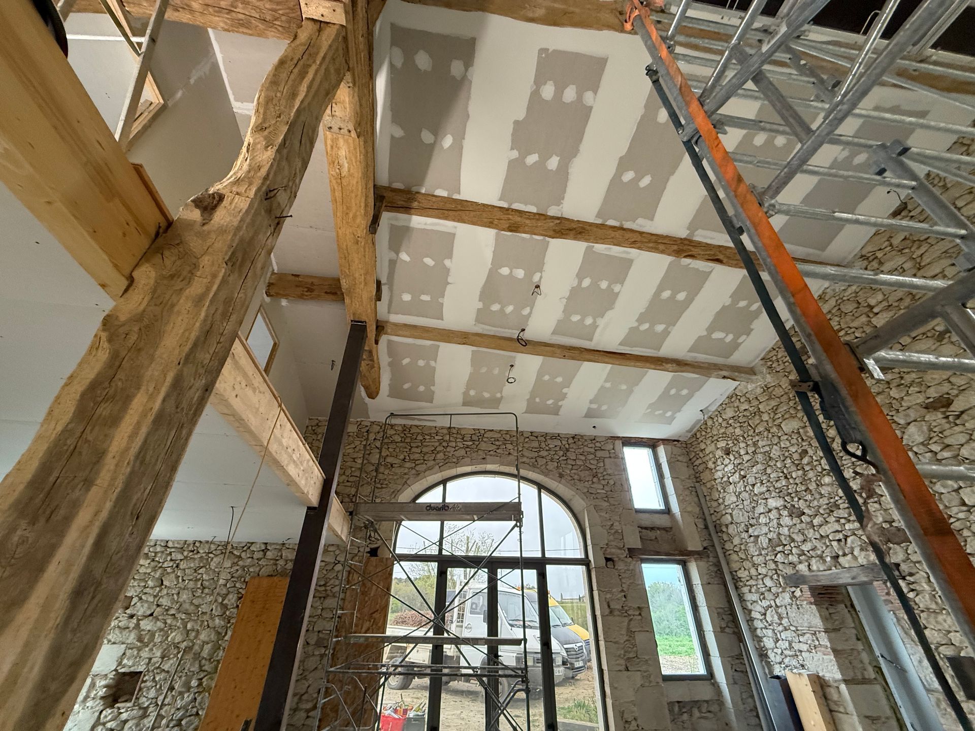 Intérieur en travaux avec poutres apparentes, plafond en plaques de plâtre, échafaudages et une grande fenêtre cintrée.