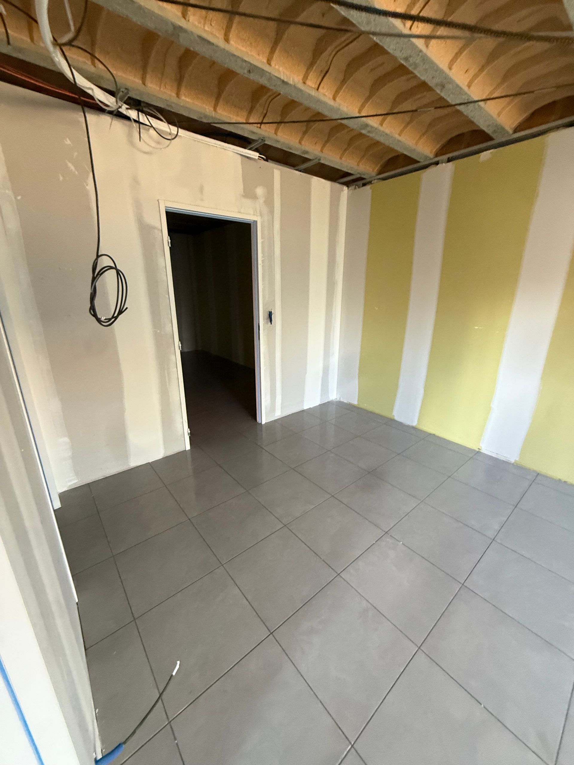 Pièce vide avec sol carrelé gris, plafond apparent et mur rayé beige et blanc.
