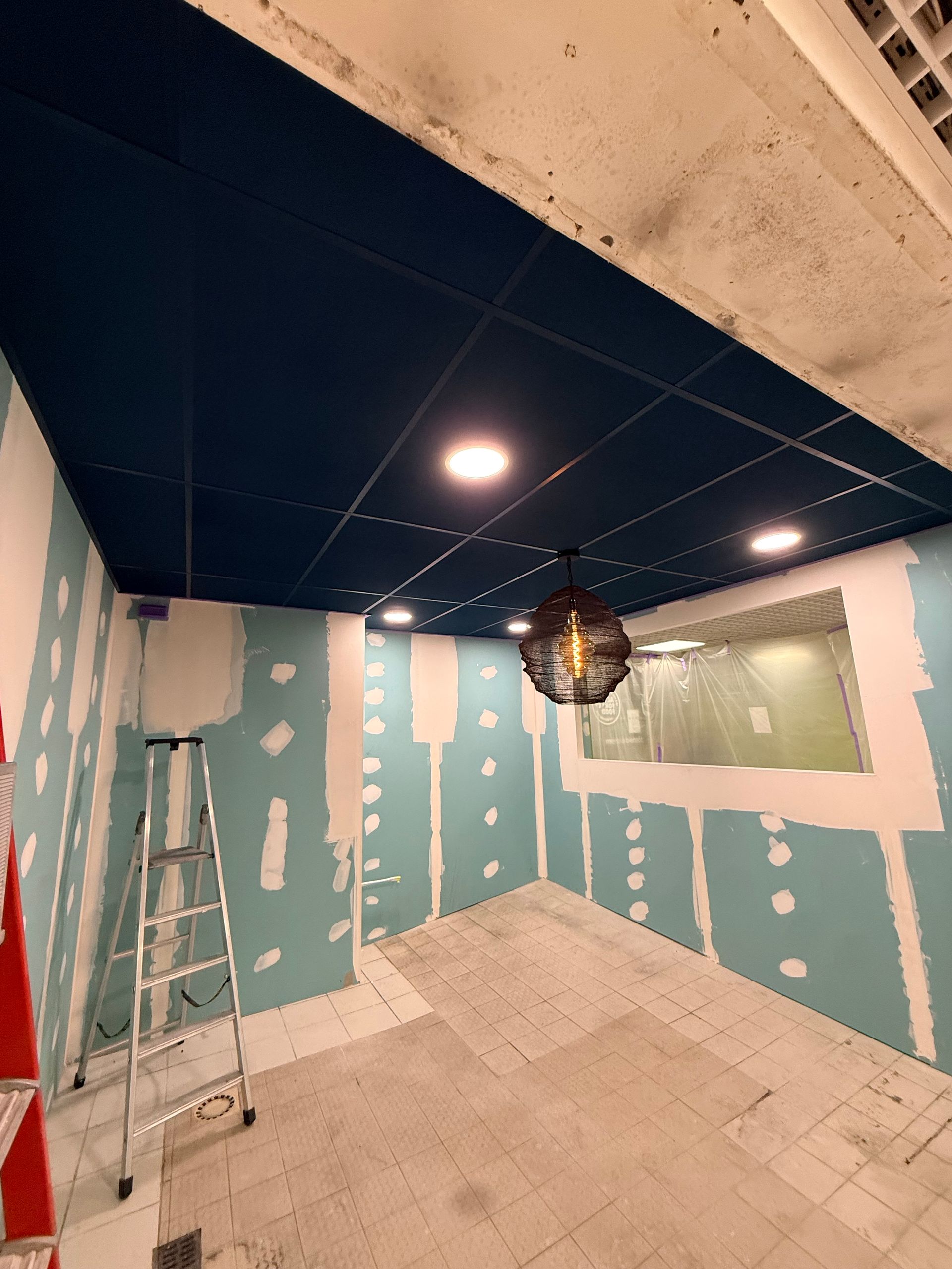 Pièce inachevée avec des plaques de plâtre bleues, un sol carrelé blanc et un faux plafond noir avec des lumières.