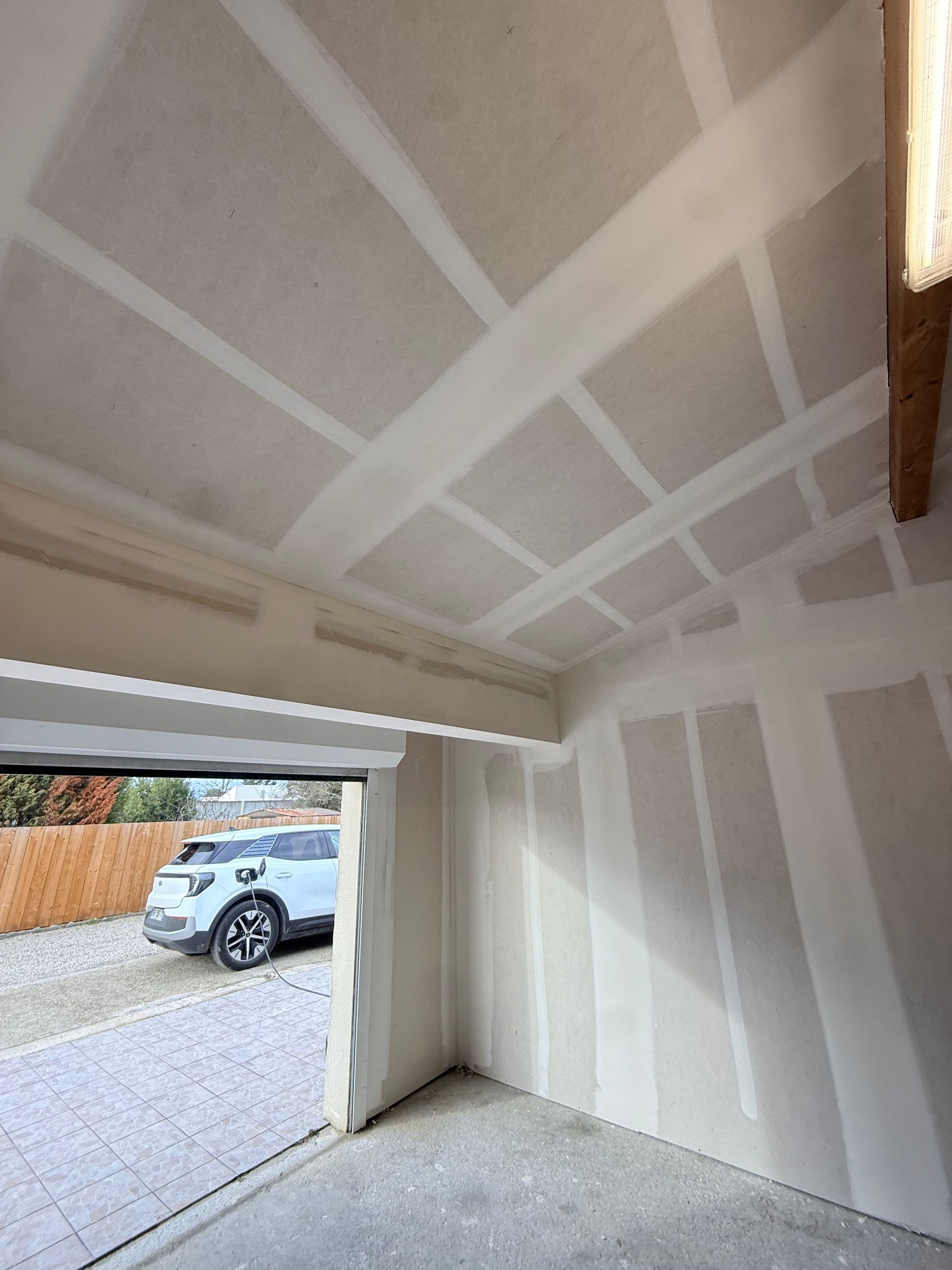 Intérieur d'un garage vide avec cloisons blanches, porte ouverte et voiture garée visible à l'extérieur.