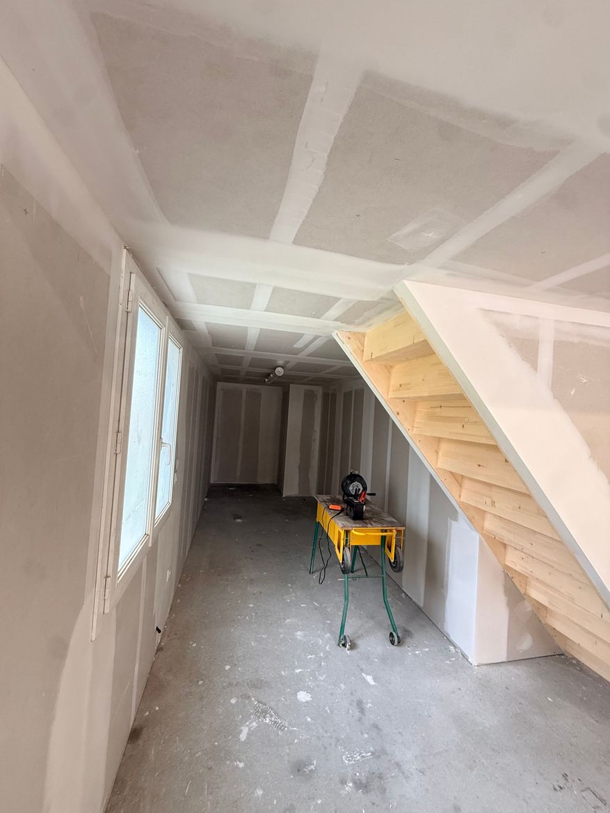 Pièce étroite et inachevée avec cloisons sèches, joints de plafond apparents et support de scie à onglets jaune.