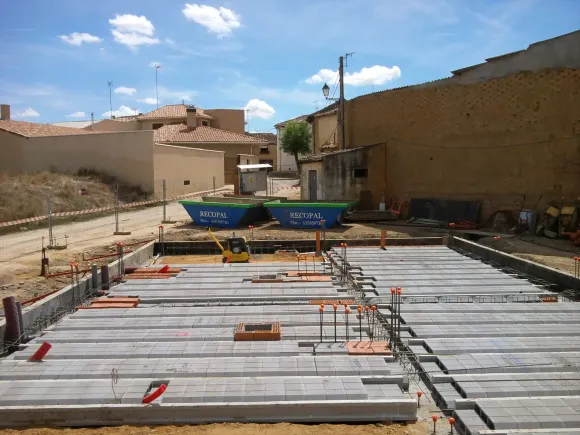 Sitio de construcción con bloques de hormigón, contenedores de basura azules y edificios bajo un cielo nublado.