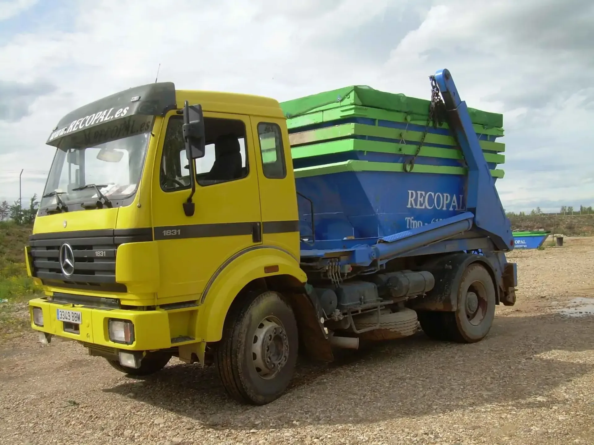 Camión volquete Mercedes amarillo y azul sobre grava, cargado con contenedores verdes y azules.