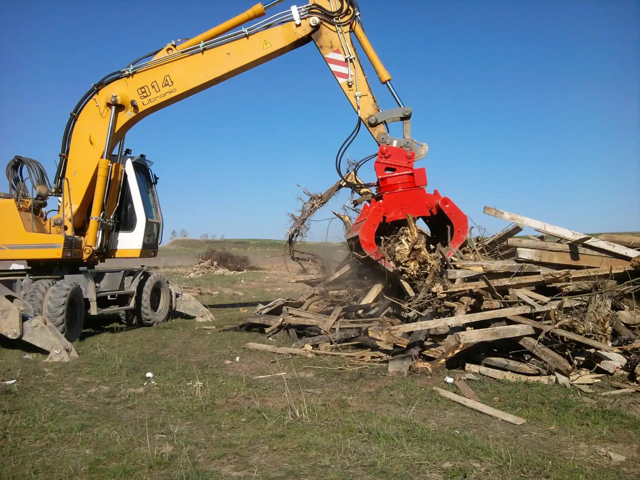 Excavadora amarilla con brazo de agarre rojo levantando escombros de madera en un campo de hierba bajo un cielo azul.