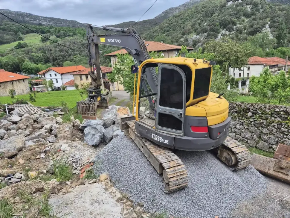 Excavadora amarilla sobre grava en un entorno rural, cerca de casas de piedra y colinas verdes.