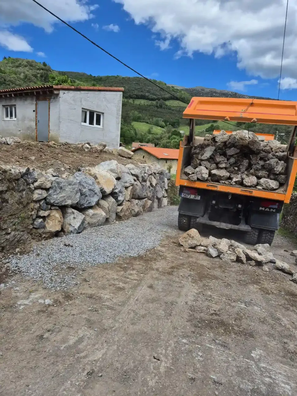 Camión volquete naranja cargado de rocas en un camino de tierra junto