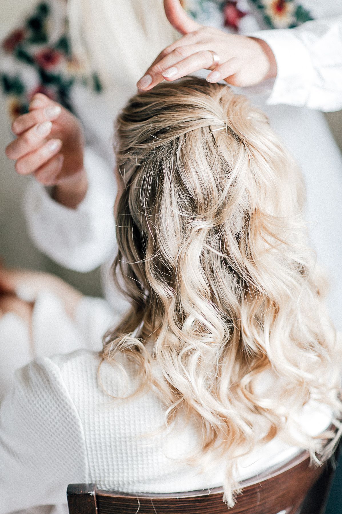 Eine Stylistin stylt blondes, gelocktes Haar zu einer halboffenen Frisur, wobei ihre Hände beim Frisieren sichtbar sind.