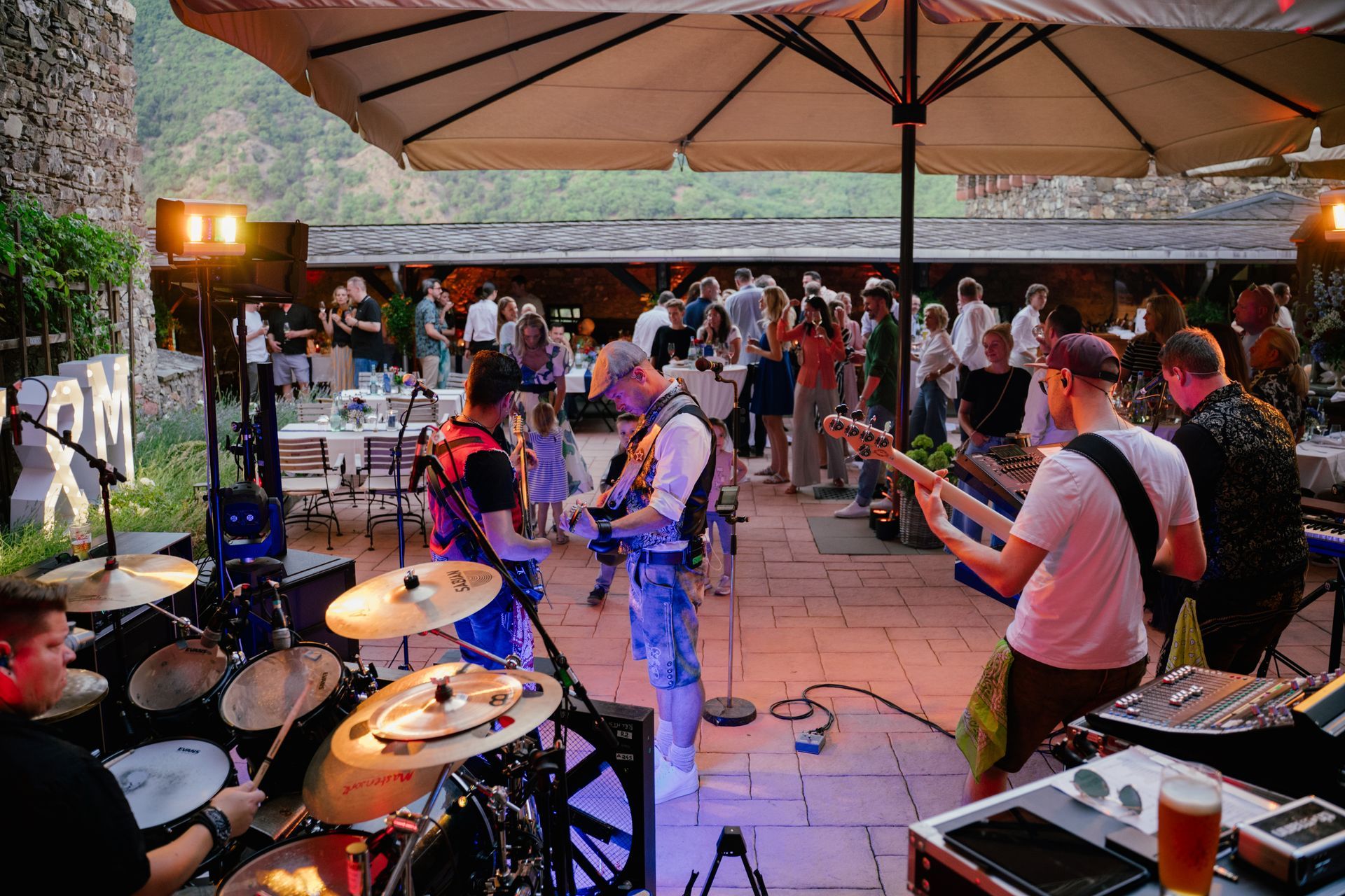 On a stone terrace under a large umbrella, a live band plays music while guests dance and dine in the background.